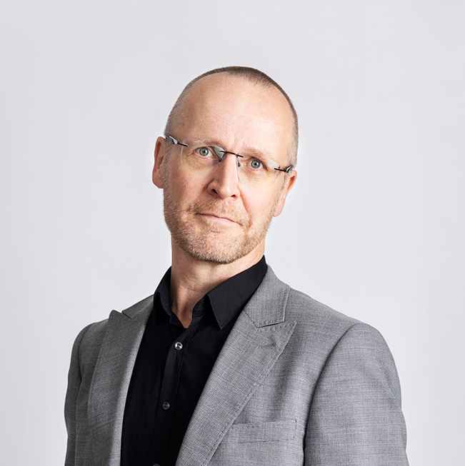 A portait of Jussi Lassila with a neutral expression, wearing a grey suit with black shirt underneath, the top button undone. He is standing slightly sideways to the camera.