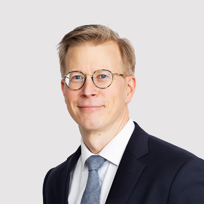 Teemu Tammikko, dark suit, light blue tie, glasses, light brown hair.