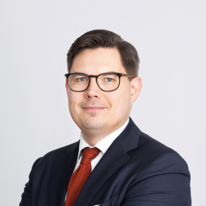 Matti Pesu, blue suit, red tie, glasses, dark brown short hair.