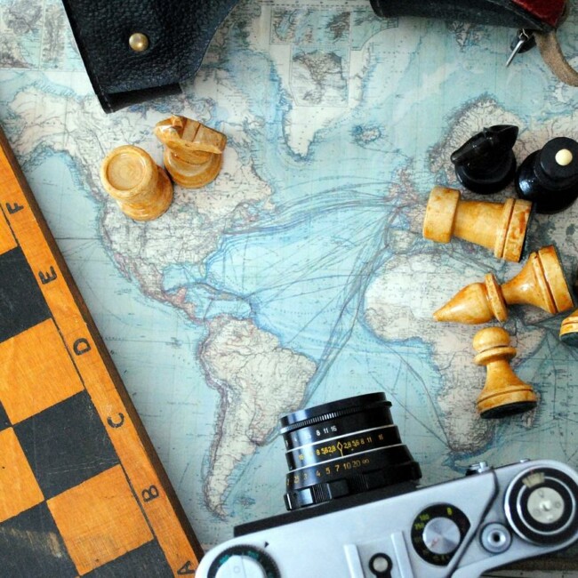 A corner of a chessboard with scattered chess pieces, a vintage camera, and a world map in the background.