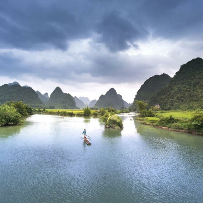 A serene river with a single boat, surrounded by lush greenery and karst mountains under a cloudy sky