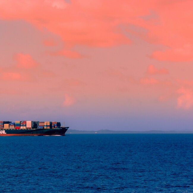 A large cargo ship with colorful containers sailing on a calm sea under a pink and orange sky.