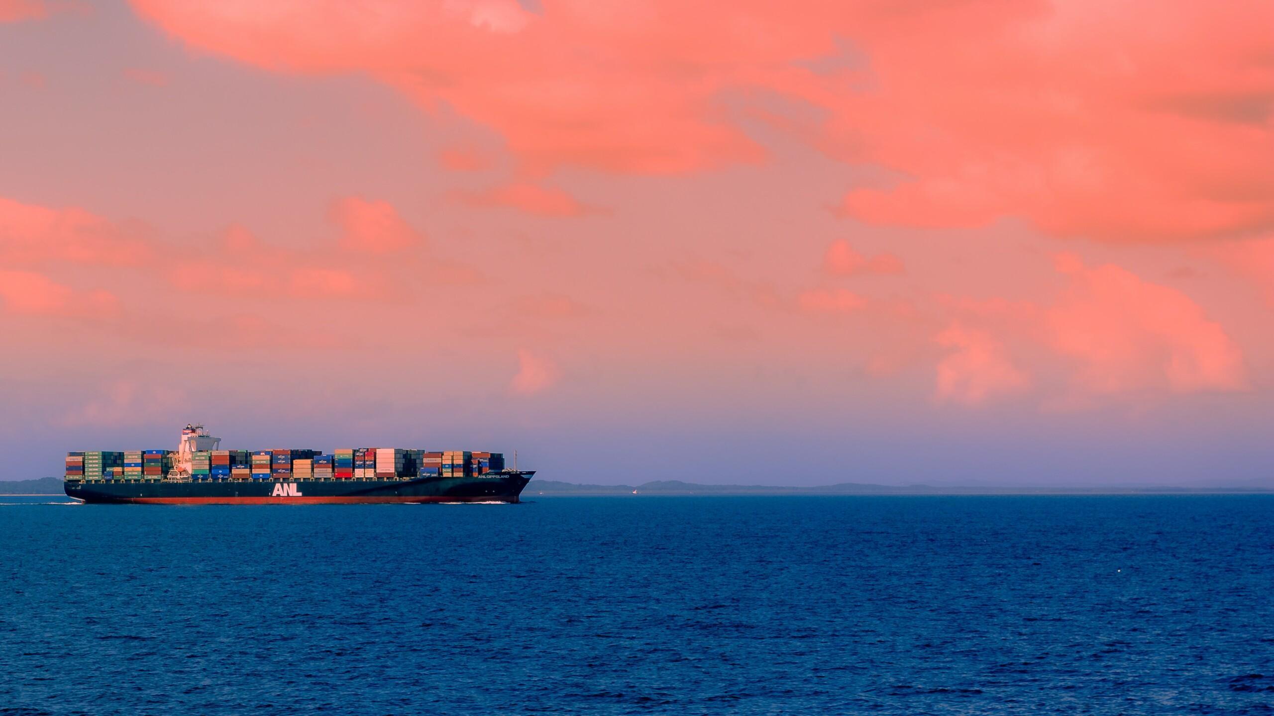 A large cargo ship with colorful containers sailing on a calm sea under a pink and orange sky.