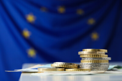 A stack of coins and banknotes and the EU flag behind them.