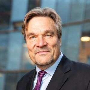 Kimmo Kiljunen with a black suit on with a purple tie. Glass wall in the background.