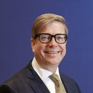 Jukka Salovaara looking at the camera with a blue suit on, blue background.