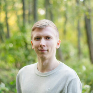 Hannes Koljonen looking at the camera in a green forest. Blonde short hair and a beige knit.