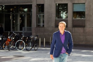 Tuomas Ojanen walking towards the camera on the street. A purple jacket and jeans on.