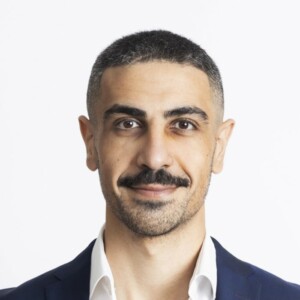 Portrait of Mohammed Hadi with a slight smile, wearing a navy suit jacket, a white dress shirt with the top button undone, and a breast pocket handkerchief. He is positioned slightly sideways to the camera.