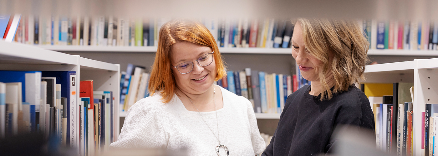 Two women in the library reading a book. Other with brown hair, other has glasses.