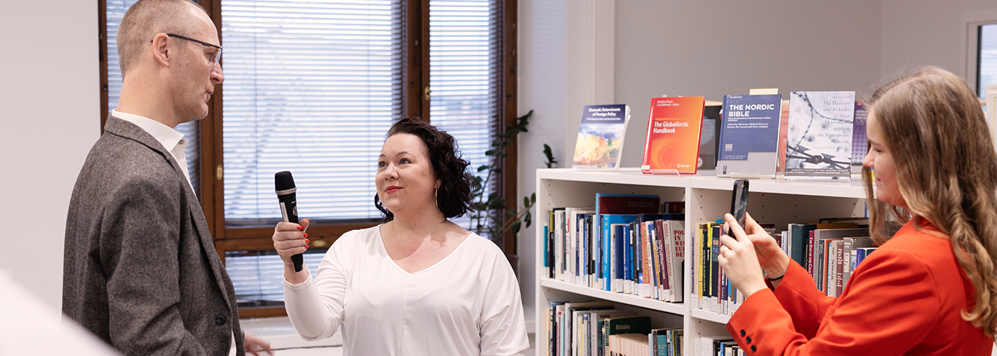 Fiia's library, where a woman with black hair and white shirt is interviewing researcher who is wearing brown suit. A woman with red suit and brown hair is filming the interview.