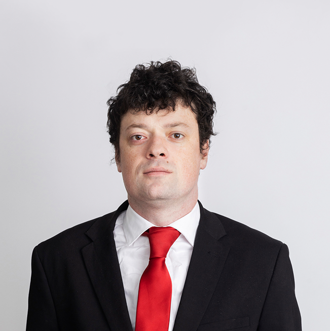 Eoin McNamara looking at the camera. He has short and curly dark hair. He is wearing a white shirt with a red tie and a black suit.