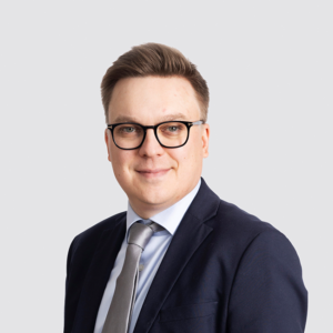 A portrait of Ville Sinkkonen with a neutral expression, wearing a dark navy suit, a light blue button-up with a silvergrey tie. He is standing slightly sideways to the camera.