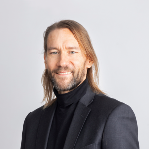 Toni Alaranta looking at the camera and smiling. He has a long brown hair and he is wearing a black polo shirt and a dark grey suit.