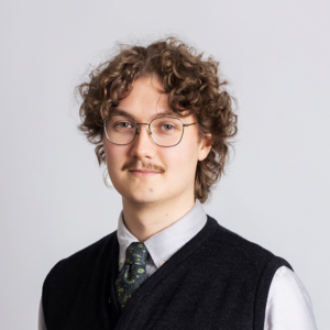 Picture of Tomi Kristeri, a slight smile on his face, glasses, dark brown hair, light grey shirt with a dark tie and a vest on top.