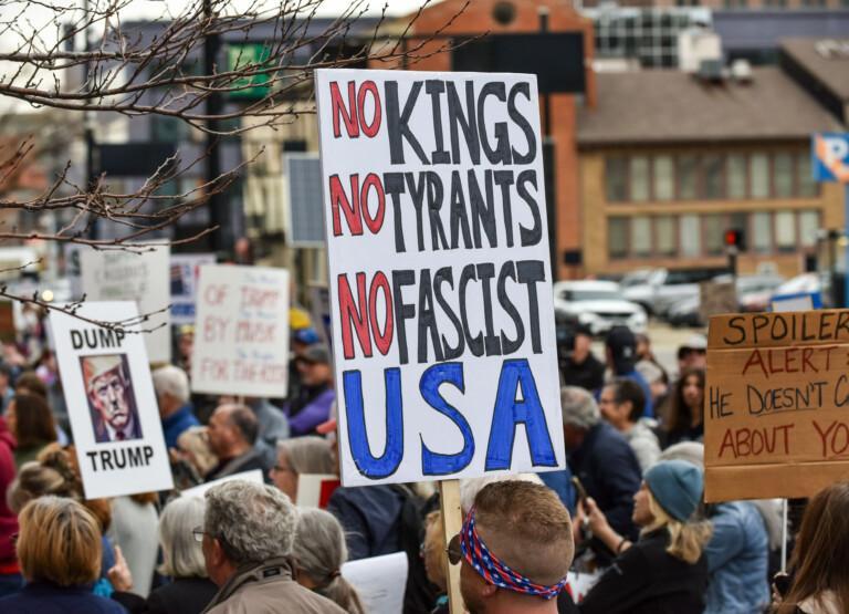 Protesters hold signs at a demonstration, including one reading No kings, no tyrants, no fascist USA.