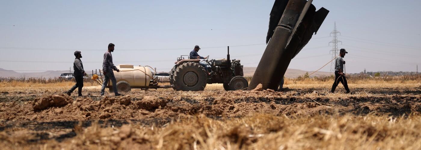 A dry field with three men and a tractor, next to a large unexploded artillery shell embedded in the ground.