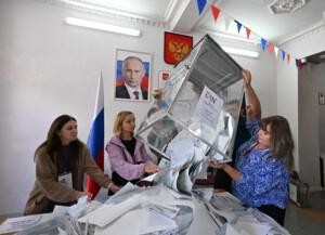 Five people gathered around a table emptying a ballot box, with a portrait of President Putin and the Russian flag in the background.