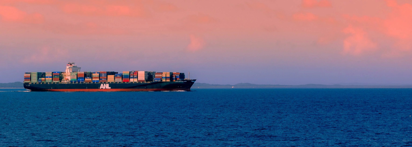 A large cargo ship with colorful containers sailing on a calm sea under a pink and orange sky.